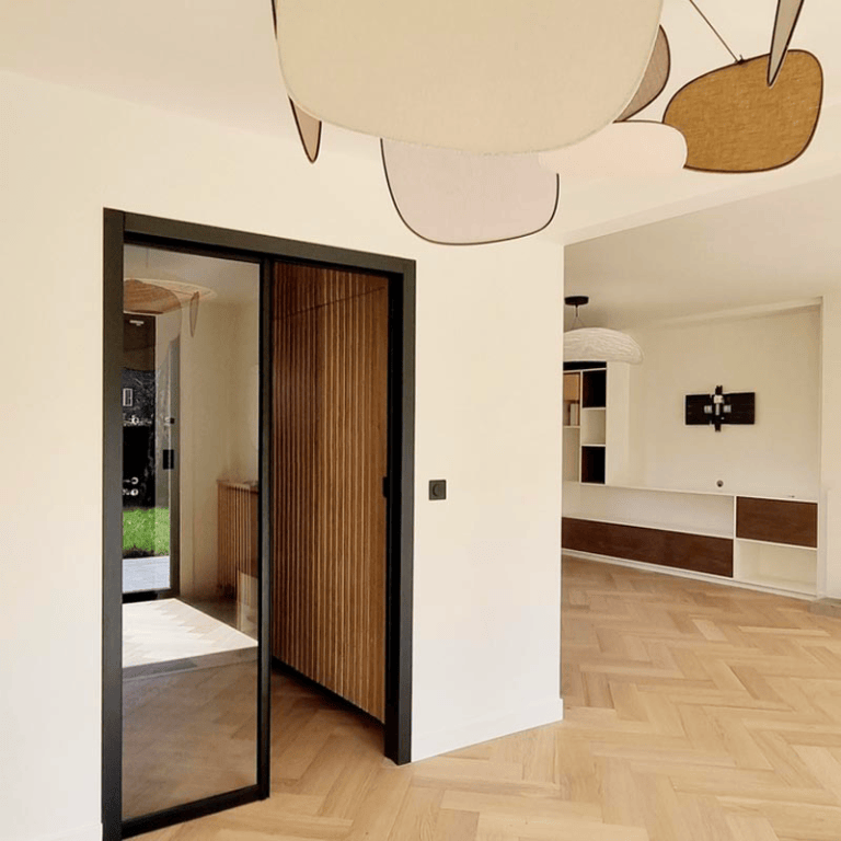 Un espace de vie lumineux avec un sol en parquet à chevrons, une porte vitrée noire et une décoration intérieure épurée.